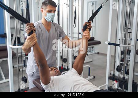 Männlicher Patient, der an Fitnessgeräten arbeitet, die von einem Physiotherapeuten überwacht werden Stockfoto