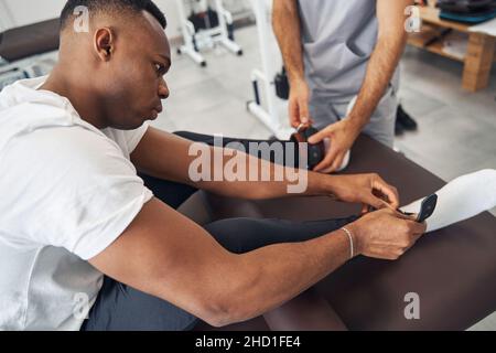 Mann, der Gewichte an seine Knöchel legte, unterstützt von einem Physiotherapeuten Stockfoto