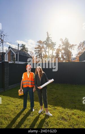 Lächelnde Frau hält eine Rolle mit Blaupausen neben einem männlichen Hausbauer, der im Hof steht und nach oben schaut Stockfoto
