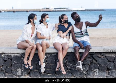 Eine Gruppe verschiedener Freunde in Masken sitzt an der Steingrenze in der Nähe des Strandes und nimmt Selfie auf dem Smartphone auf, während sie das Wochenende auf Lanzarote genießen Stockfoto