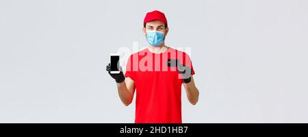 Kontaktlose Lieferung, Bezahlung und Online-Shopping während covid-19, self-Quarantäne. Lächelnder Kurier in roter Uniform, Handschuhe und Gesichtsmaske zeigen smart Stockfoto