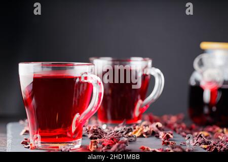 Hibiskus-Tee in Glaskrug auf dunklem Hintergrund. Tasse roten Hibiskustee und trockene Hibiskusblüten auf einer Schiefertafel Stockfoto