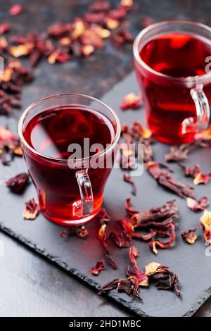 Hibiskus-Tee in Glaskrug auf dunklem Hintergrund. Tasse roten Hibiskustee und trockene Hibiskusblüten auf einer Schiefertafel Stockfoto