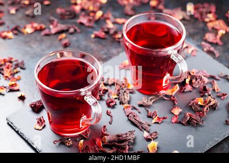 Hibiskus-Tee in Glaskrug auf dunklem Hintergrund. Tasse roten Hibiskustee und trockene Hibiskusblüten auf einer Schiefertafel Stockfoto