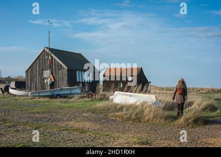 Suffolk-Küste, Rückansicht einer Frau, die allein an alten Fischerhütten am Walberswick Quay entlang der Suffolk-Küste, England, vorbeigeht Stockfoto