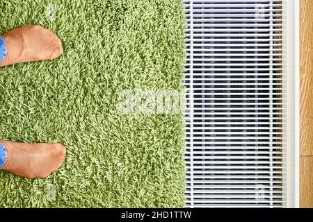 Moderner Wasser-Fußbodenwärmekonvektor in der Nähe des Panoramafensters mit grünem Teppich und Füßen in orangefarbenen Socken aus der oberen Ansicht installiert Stockfoto