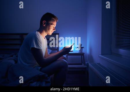 Einsamer Mann, der im Schlafzimmer auf dem Bett saß und nachts telefoniert. Gesicht vom Smartphone-Display beleuchtet. Stockfoto