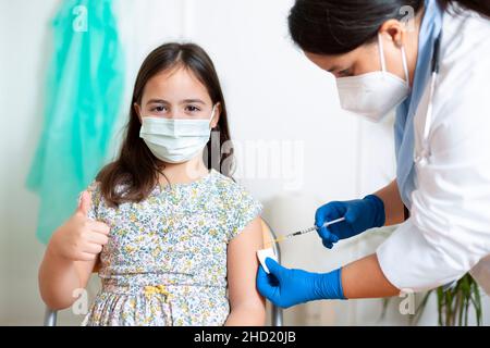 Kleines kaukasisches Kind, das einen Daumen nach oben gibt, während es von einer medizinischen Mitarbeiterin geimpft wird. Impfkampagne gegen Kinder. Stockfoto