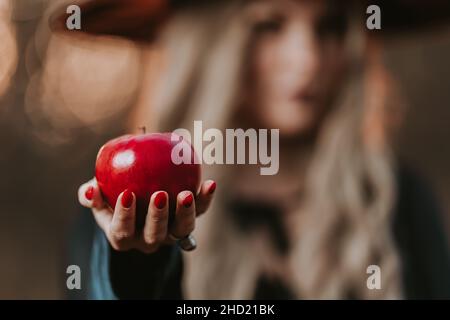 Hexe Frau in schwarz bietet roten Apfel als Symbol der Versuchung, Gift. Märchenhaftes Konzept des weißen Schneeassistenten. Gruseliges halloween, Cosplay. Stockfoto