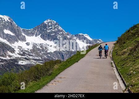 Zwei Radler mit E-Bikes fahren in der Ferne das Wienwasserntal, schneebedeckte Berge oberhalb von Andermatt hinauf. Stockfoto