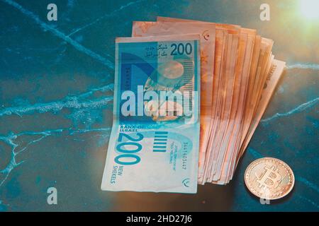 Rollen Sie israelische Schekel ILS-Banknoten in der Nähe von Kryptowährungstoken Bitcoins Bitcoins, von oben gesehen, auf einem alten Marmorbrett. Selektiver Fokus. Mit Platz für die Textplatzierung. Hochwertige Fotos Stockfoto