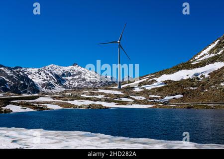 Eine Windturbine, gesehen über einen See, auf dem Gotthard Pass auf 2106 m, umliegender schneebedeckter Berge. Stockfoto