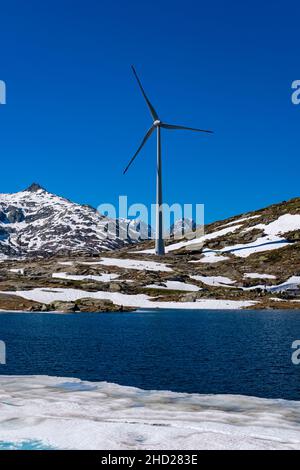 Eine Windturbine, gesehen über einen See, auf dem Gotthard Pass auf 2106 m, umliegender schneebedeckter Berge. Stockfoto