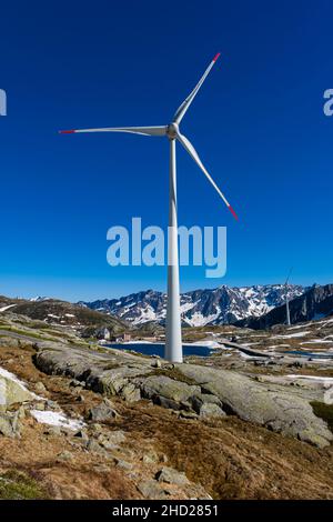 Eine Windturbine, gesehen über einen See, auf dem Gotthard Pass auf 2106 m, umliegender schneebedeckter Berge. Stockfoto