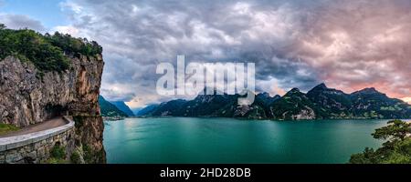 Panoramablick auf den Sonnenuntergang über dem Vierwaldstättersee, die Stadt Flüelen in der Ferne am Ufer, hohe Berge über dem See. Stockfoto