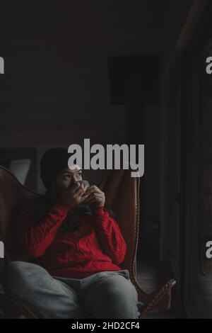 Vertikale Aufnahme einer hispanischen Frau in rotem Pullover und Trinken aus einem Becher auf ihrem Schmetterlingsstuhl Stockfoto