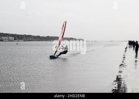 Ein Windsurfer nutzt einen windigen Tag, an dem er mit hoher Geschwindigkeit an Wanderern am Rand des Marine Lake in West Kirby auf der Wirral Peninsula vorbeikommt Stockfoto