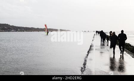 Walkers am Neujahrstag 2022 Wandern am Rande des Marine Lake in West Kirby auf dem Wirral, während ein Windsurfer die windigen Bedingungen genießt. Stockfoto