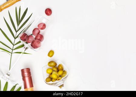 Mediterrane Lebensmittelkomposition mit Weingläsern mit Trauben und grünen Oliven, Flasche Rosenwein und Palmenblättern auf weißem Hintergrund mit Kopierraum Stockfoto