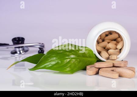 Weiße Botle mit Kräutertabletten, Stethoskop und grünen Blättern auf hellblauem Hintergrund aus nächster Nähe. Konzept der Kräutermedizin Stockfoto