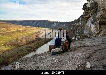 Junger Mann mit Mädchen sitzt auf dem Rand der Klippe mit Beinen baumeln und Blick auf den Fluss. Mann hat Frisur mit Dreadlocks. In der Nähe des Felsenklosters, Stockfoto