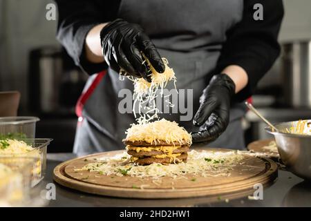 Der Koch bereitet in der Küche Leberkuchen zu. Nahaufnahme der weiblichen Hände in Handschuhen mit Käseleber-Pfannkuchen bestreuen. Stockfoto