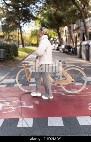 Seitenansicht einer Frau in Oberbekleidung, die an sonnigen Tagen in der Stadt Fahrrad schiebt und auf einem Pfad über eine Asphaltstraße läuft. Stockfoto