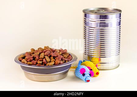 Braune knusprige Bio-Kitty-Kibble-Stücke in einer Schüssel, Spielzeugmäuse und nasses Katzenfutter aus Metalldose für Tierfutter auf hellem Hintergrund. Stockfoto