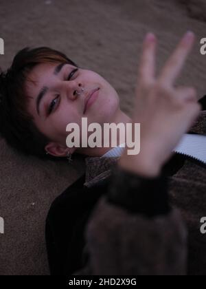 Nicht-binäre Frau, die mit dem Kopf auf dem Sand liegt und mit der Hand ein V-Zeichen macht Stockfoto