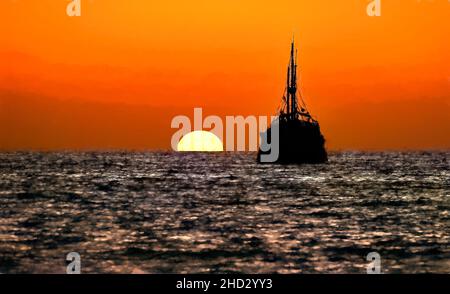 Piratenschiff Ocean Sunset Silhouette ist ein altes hölzernes Piratenschiff mit vollen Flaggen, wenn die Sonne am Ozeanhorizont in einem bunten Sonnenuntergangshimmel untergeht Illustr Stockfoto