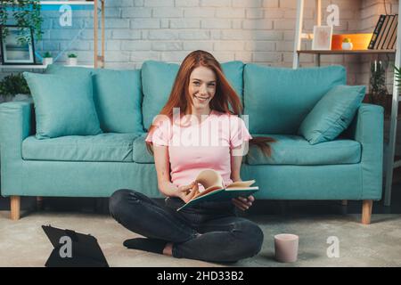 Kaukasische Frau, die zu Hause studiert, auf dem Boden sitzt und Tagebuch liest. Rotkopffrau. Lächelndes Gesicht. Quarantäne-Lebensstil. Coronavirus Stockfoto