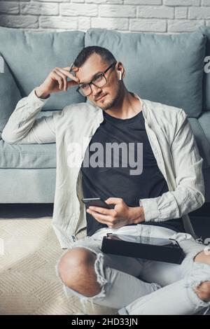 Vorderansicht des kreuzbeinigen Mannes, der während des COVID-19 mit dem Telefon und Tablet auf dem Boden sitzt. Stockfoto