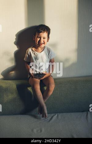 Kaukasischer kleiner Junge lächelt der Kamera zu, während er eine Schale mit Blaubeeren isst, die zu Hause auf der Couch sitzt. Stockfoto
