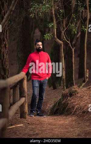 Vertikale Aufnahme eines bärtigen hispanischen Mannes in einem roten Pullover im Wald Stockfoto