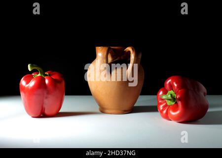 Tontopf und zwei rote Paprika auf beiden Seiten auf einer weißen Oberfläche mit schwarzem Hintergrund Stockfoto