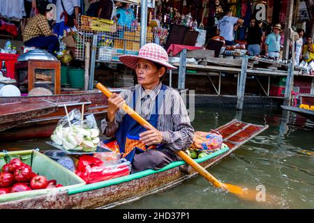 Thailändische Dame Bootsfahrerin mit Essen in ihrem Boot rudert an einer Gruppe von Menschen im Hintergrund Land Teil des Marktes vorbei. Stockfoto