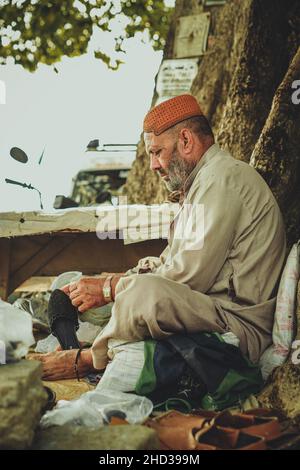 Armer alter Schuhmacher, der mit seinen Werkzeugen unter einem Baum in einem lokalen Straßenladen Pakistans sitzt Stockfoto