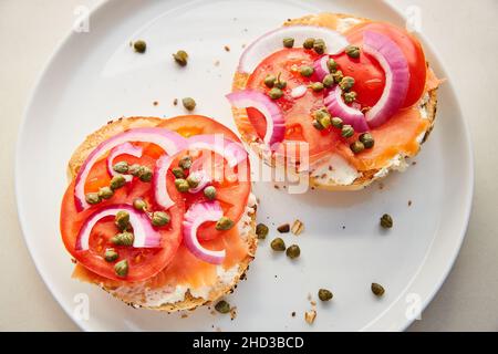 Draufsicht auf Lox, Tomaten, Kapern, rote Zwiebeln und Frischkäse auf Bagel auf weißem Teller Stockfoto