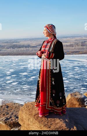 Im Winter steht eine junge Frau am Rand einer Klippe, gekleidet in ein nationales Baschkir-Kostüm. Stockfoto