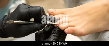 Nagelhautentfernung an den Zehen. Hände in schwarzen Handschuhen des Pediküre-Meisters entfernen die Nagelhaut an den weiblichen Zehen durch Stopfer Stockfoto
