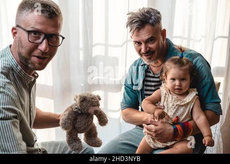 Schwule Männer mit adoptierten Mädchen zu Hause - zwei gutaussehende Väter spielen mit ihrer Tochter - LGBTQ+ Familie zu Hause - Diversity-Konzept und LGBT Stockfoto