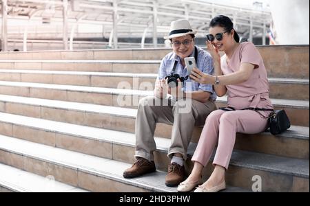 Ältere asiatische Paare sitzen auf der Treppe während der Reise und haben Spaß Videoanrufe mit jemandem. Reisekonzept für ältere Paare. Stockfoto