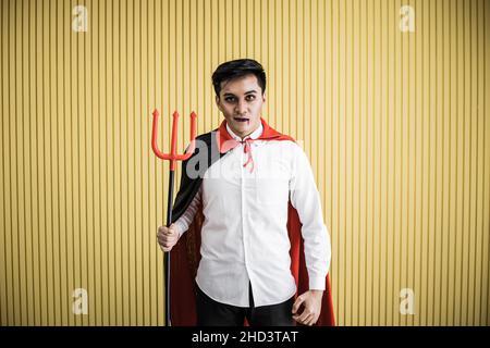 Halloween-Konzept des jungen asiatischen Mannes im Kostüm dracula und halten Speer auf gelbem Hintergrund. Porträt eines Teenagers, der zum Feiern als dracula verkleidet ist Stockfoto
