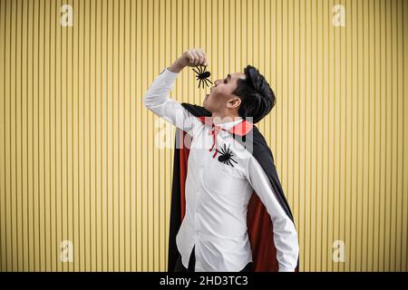 Junger asiatischer Mann im Kostüm dracula und essen schwarze Spinne auf gelbem Hintergrund von Halloween-Konzept. Porträt eines Teenagers, der als dracula für Cele verkleidet ist Stockfoto