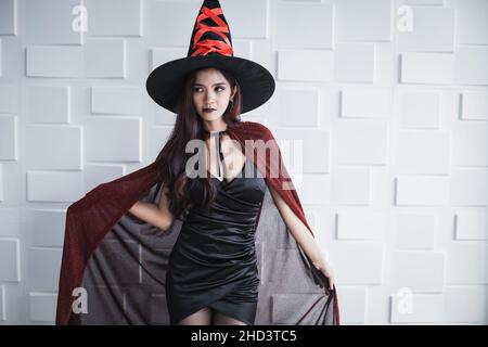 Junge asiatische Frau in Kostüm Hexe auf weißer Wand des Halloween-Konzepts. Porträt einer Teenager-Frau, die als Hexe verkleidet ist, um das Halloween-Festival zu feiern. Stockfoto