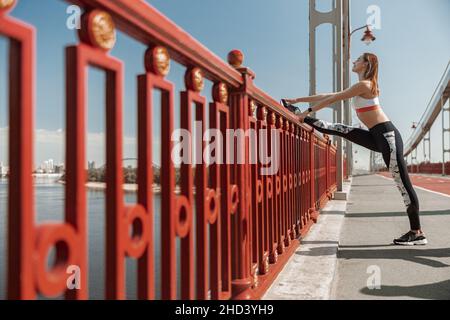 Positive Frau macht Dehnungsübungen für die Beine in der Nähe von Geländer auf Brücke Stockfoto