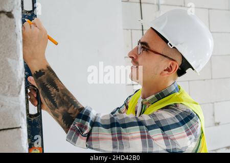 Skrupelloser Bauingenieur in einem weißen Helm, der in einem unfertigen Gebäude Markierungen an einer Ziegelmauer macht. Stockfoto