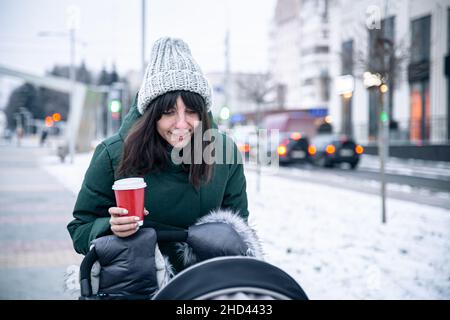 Stilvolle junge Mutter mit einer Tasse Kaffee auf einem Spaziergang mit einem Kinderwagen im Winter. Stockfoto