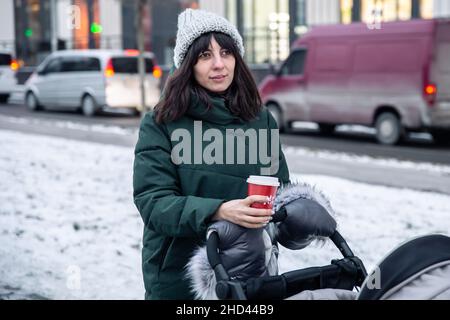 Stilvolle junge Mutter mit einer Tasse Kaffee auf einem Spaziergang mit einem Kinderwagen im Winter. Stockfoto