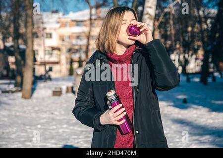 Eine junge Frau trinkt an einem sonnigen Wintertag ein heißes Getränk mit Thermoskanne. Stockfoto
